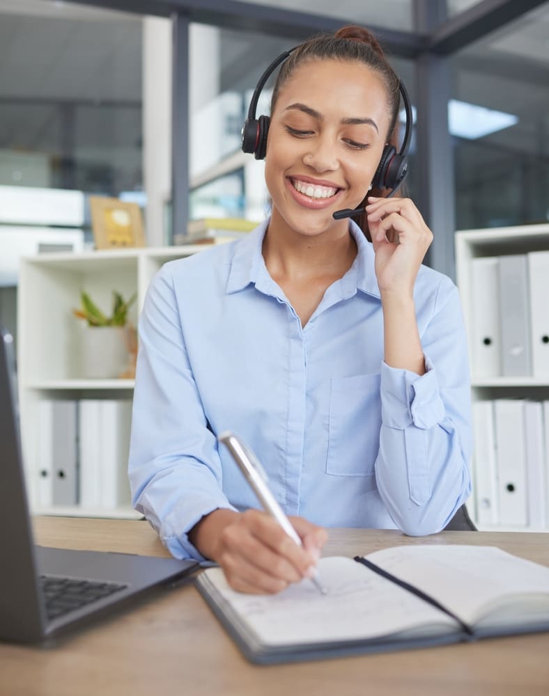 A woman with headphones and a headset smiles at her desk, working on an R&D grant.