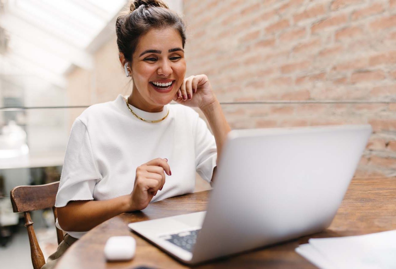 A woman sitting at a table looking at a laptop while smiling.