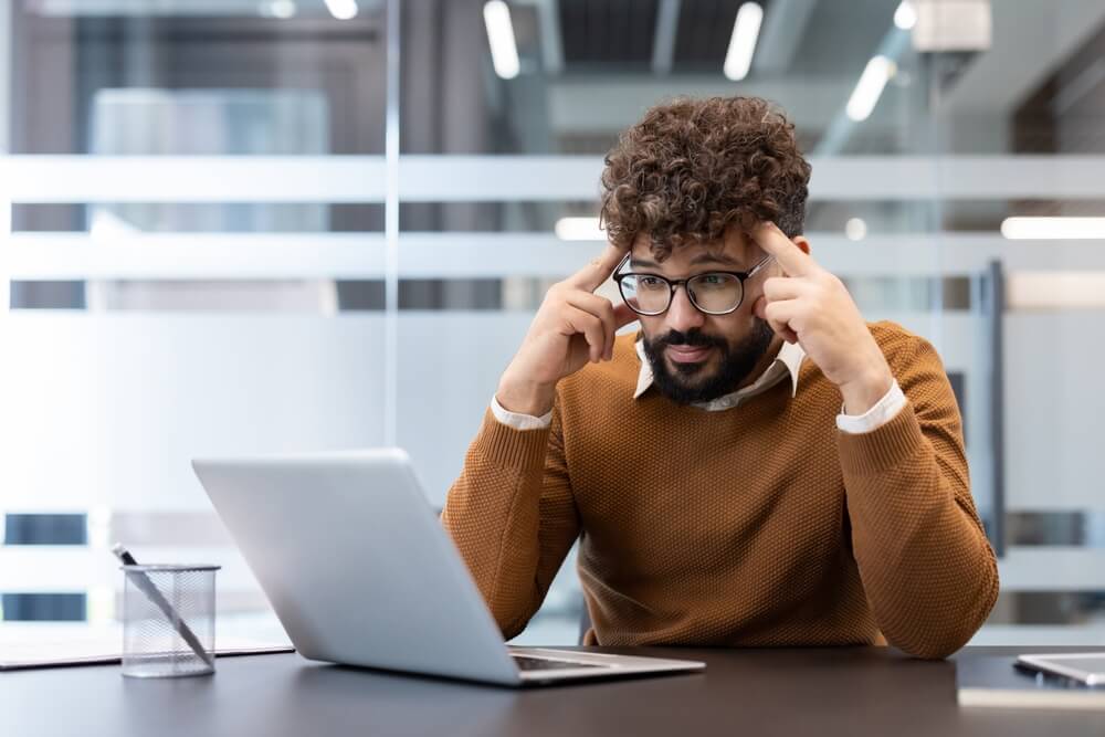 A man with glasses sitting at a laptop in an office, fingers to his temples in concentration, suggesting careful analysis or complex decision-making.