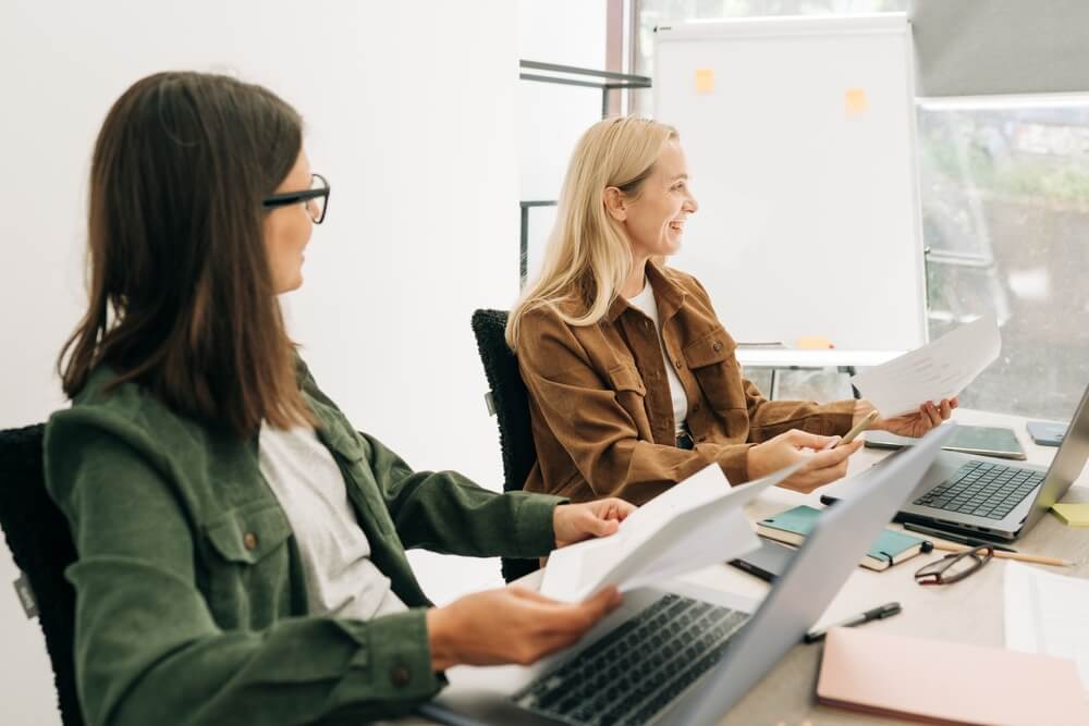 Two professionals reviewing documents together at a desk during a business advisory meeting, with laptops and a whiteboard in the background.
