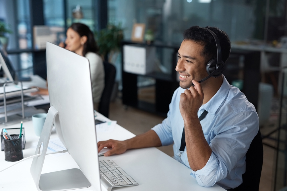 A smiling man wearing a headset works at a desktop computer in a modern open-plan office, with a colleague visible in the background.