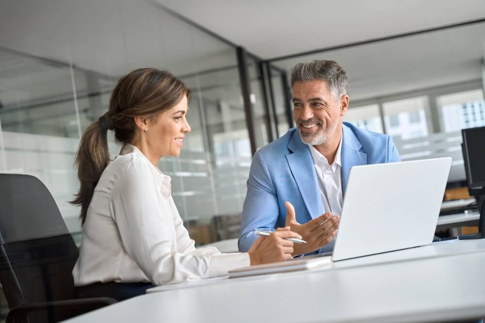 A man and woman in business attire sitting together at a desk, reviewing content on a laptop during a professional consultation.