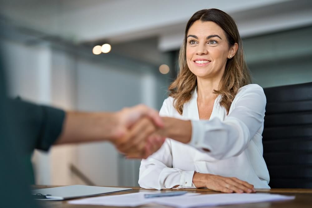 A businesswoman shaking hands across a desk in a professional office setting, suggesting a successful adviser consultation.