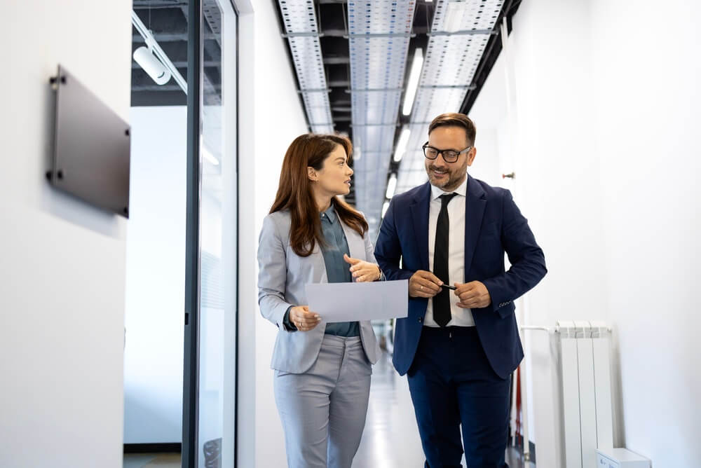 Two business professionals walking through a modern office corridor, one holding documents and gesturing during a discussion.